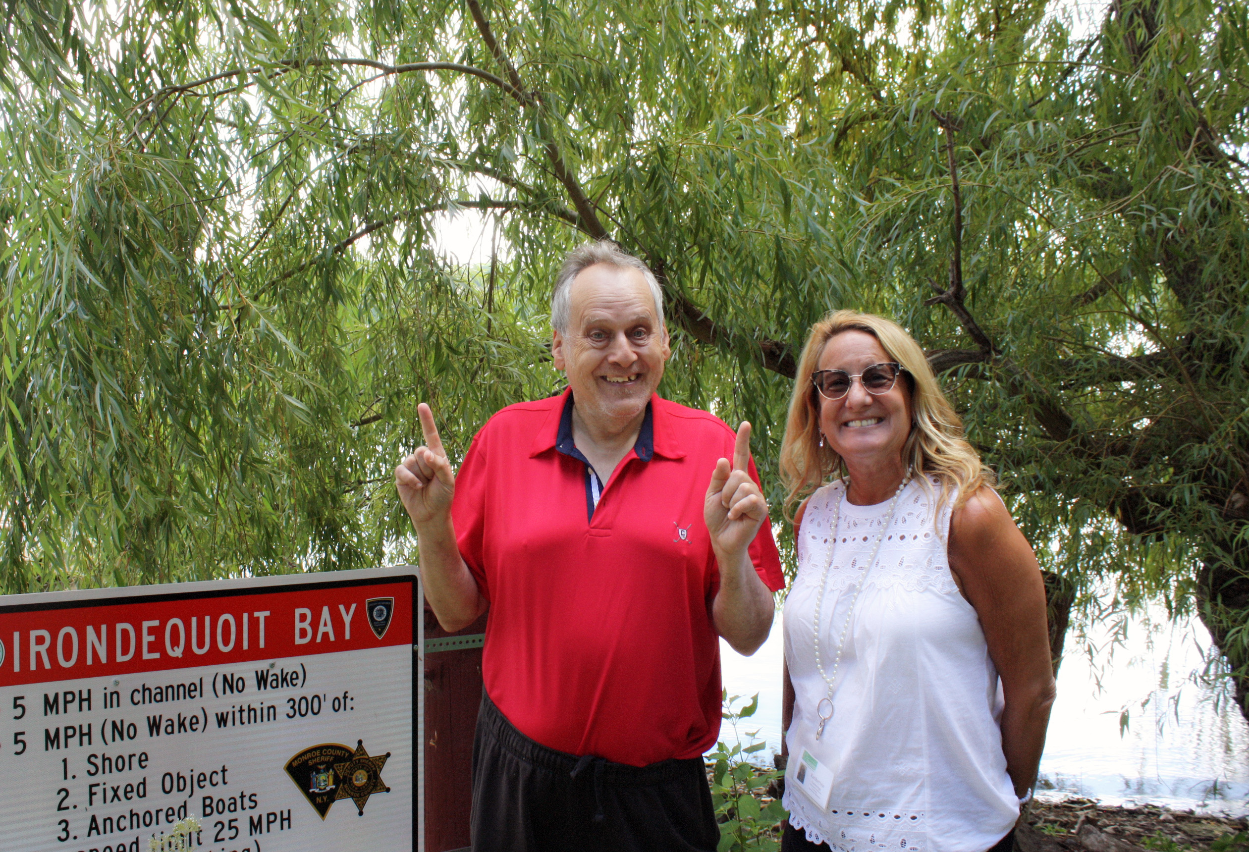 Image shows two adults smiling and standing by sign at Irondequoit Bay