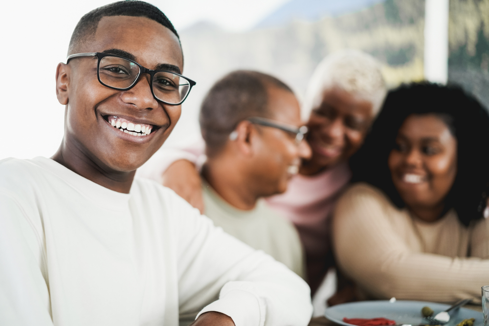 Black teen boy smiling and wearing glasses and seated at dinner table and his parents and relatives are smiling in the background